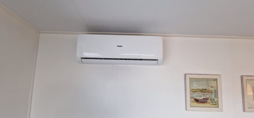 Witte airco eenheid aan de muur met twee ingelijste afbeeldingen ernaast in een lichte kamer.