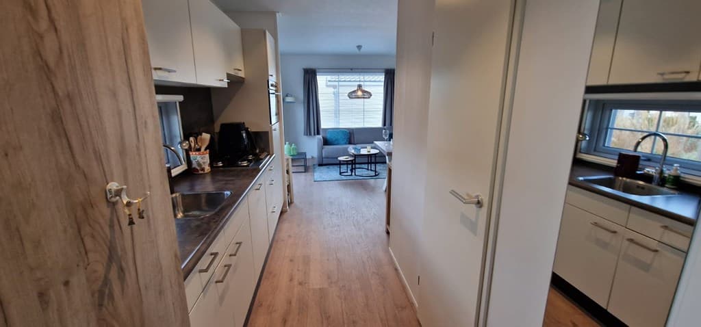 Ingericht keuken met doorgang naar een lichte woonkamer met bank en eettafel, naast een tweede keukenunit.