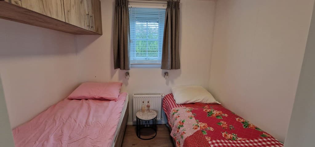 Slaapkamer met twee eenpersoonsbedden, kleurrijke dekbedden en een klein bijzettafeltje bij het raam