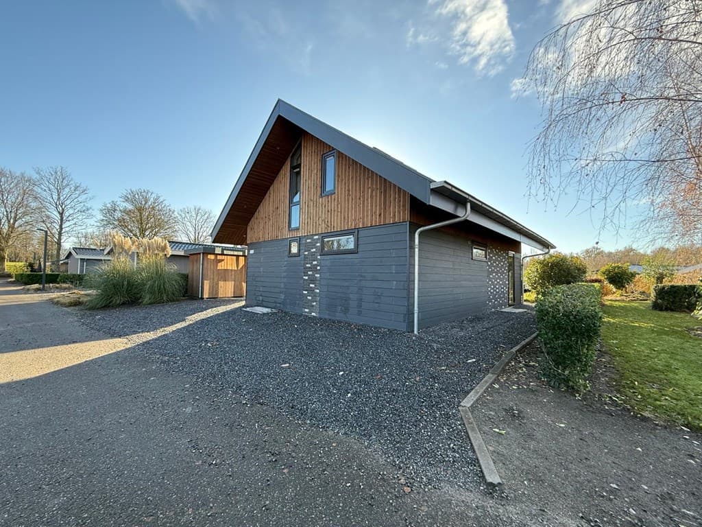 Moderne vakantiewoning met houten gevel, gelegen aan een verhard pad en omringd door groen en bomen.
