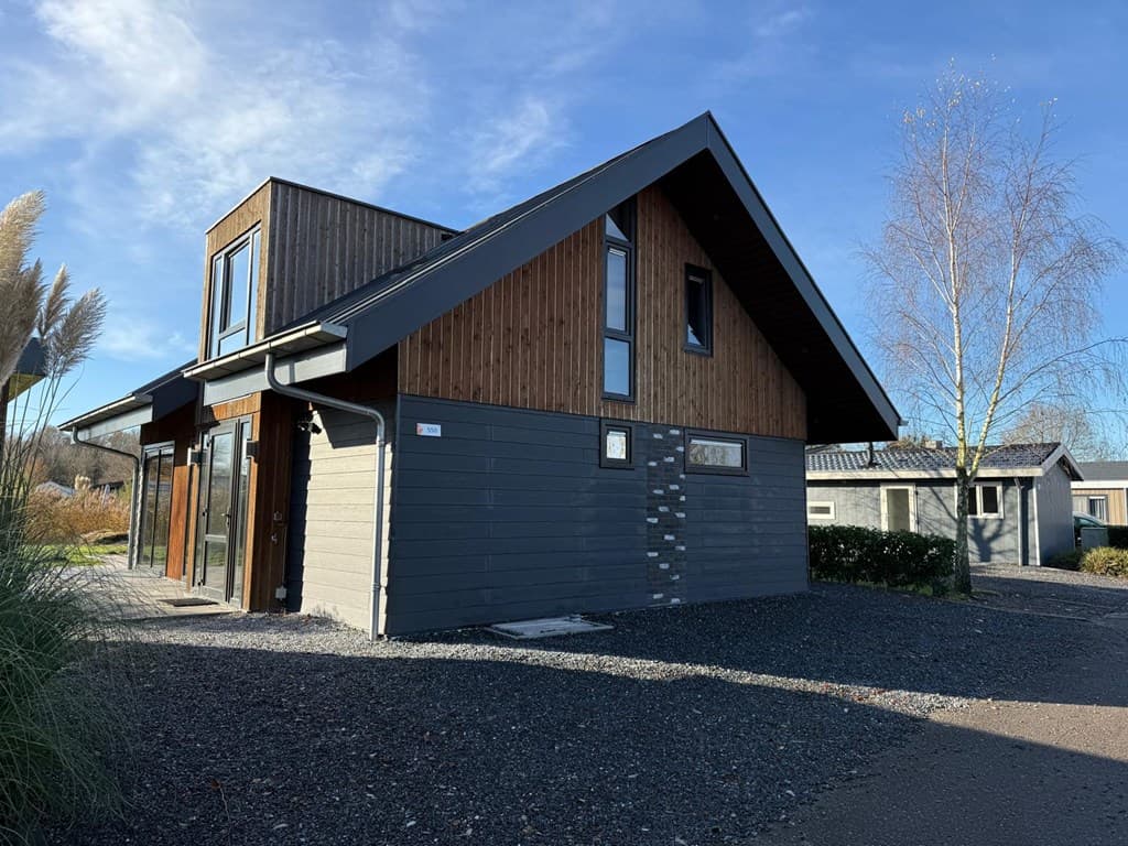Moderne vakantiewoning met houten gevel en grote ramen, omgeven door een grindpad en laurierstruiken.