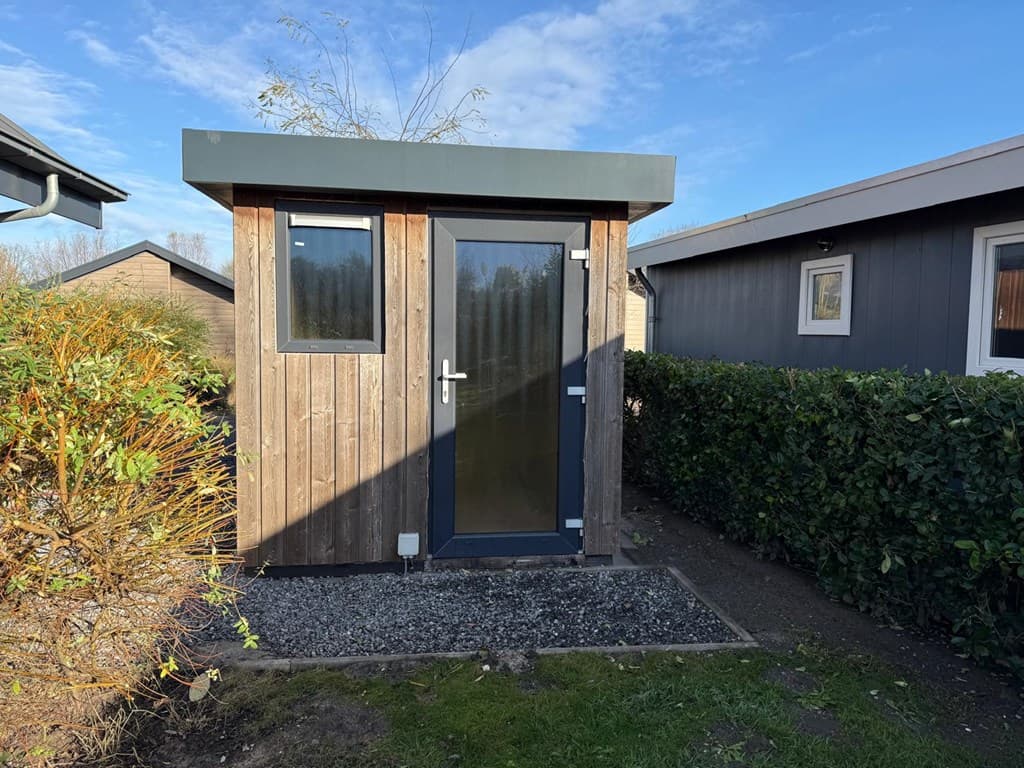 Houten tuinhuisje met een glazen deur en ramen, omringd door groene struiken en een grindpad.