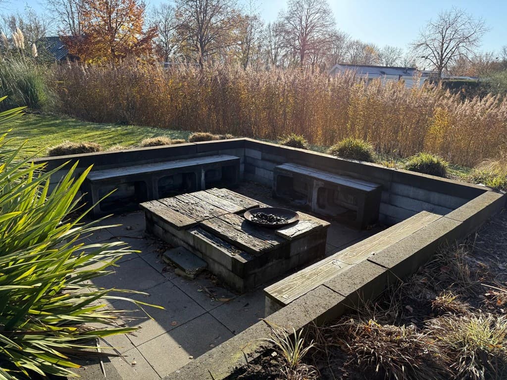 Terras met een verhoogd zitgedeelte en een vuurschaal, omringd door groen en hoge planten aan de rand van de tuin