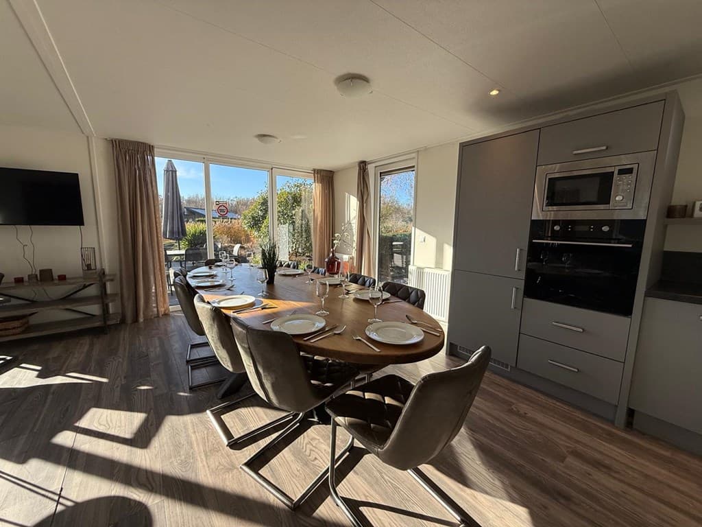 Eetkamer met eettafel en stoelen, keuken met moderne apparatuur en veel lichtinval door grote ramen.