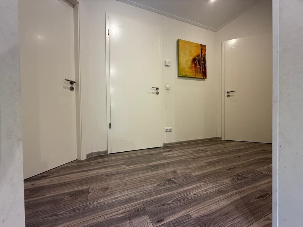 Ingang van de woning met lichtgekleurde wanden, houten vloer en deuren naar andere ruimtes.