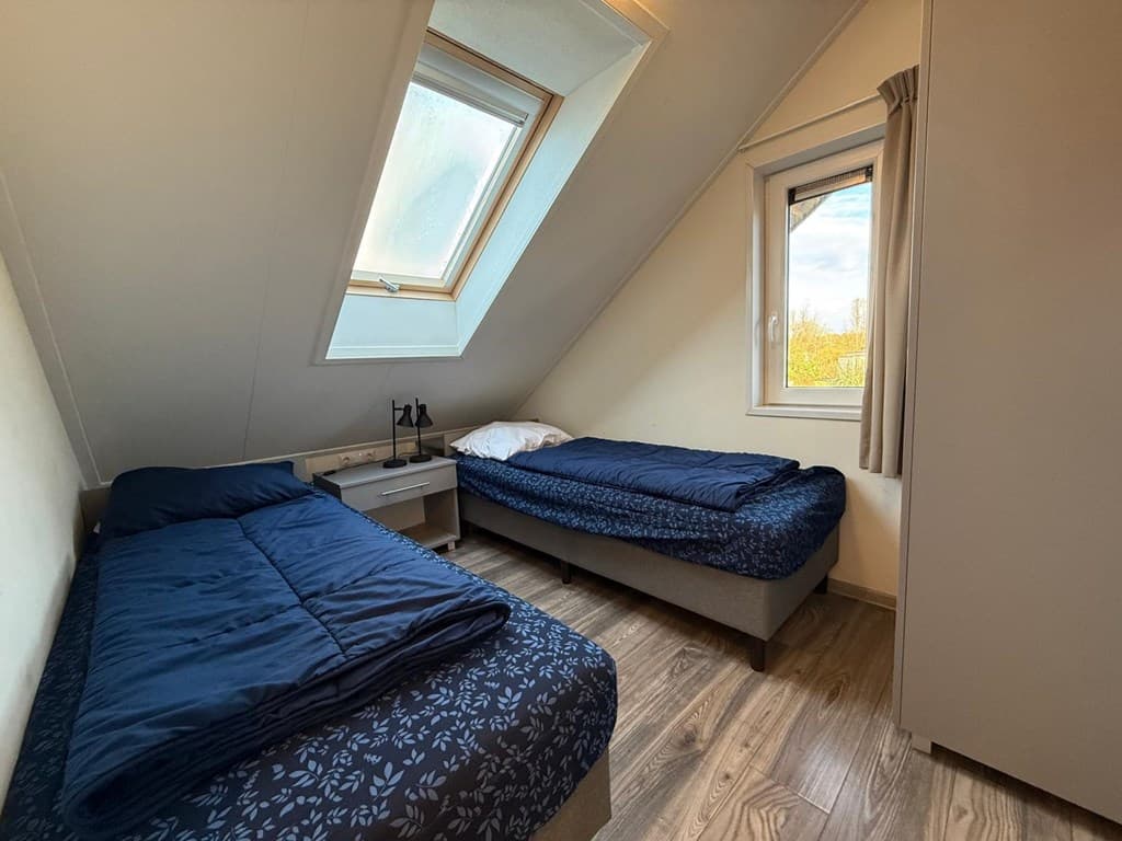 Slaapkamer met twee éénpersoonsbedden, een raam met een schuin dak en een kast in een lichte ruimte.