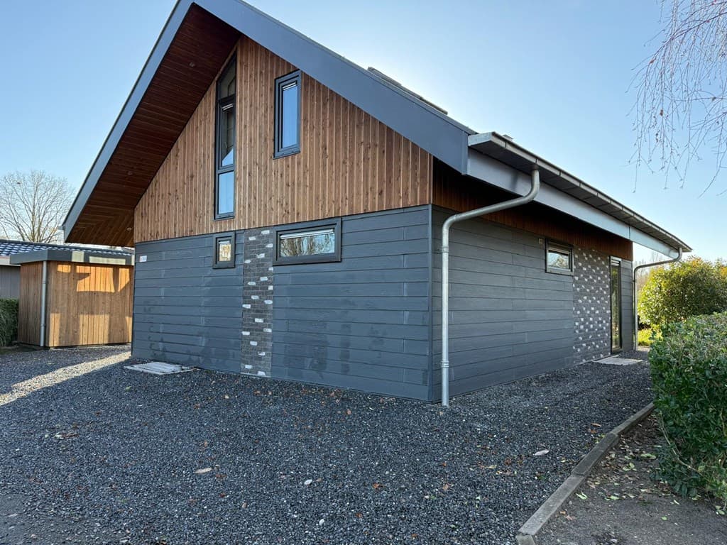 Moderne vakantiewoning met houten gevelbekleding en grijze stenen, omgeven door grind en groen.