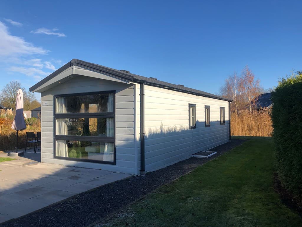 Moderne vakantiewoning met lichte houten gevel, grote ramen en een terras met stoelen en parasol in een groene omgeving