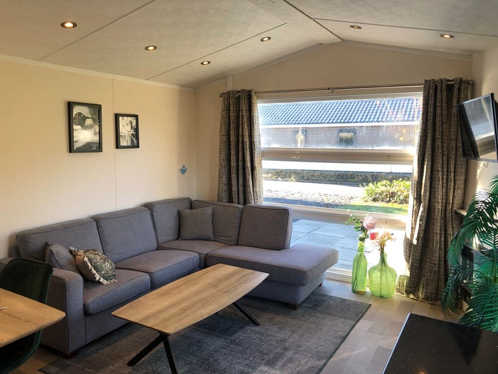 Ruime woonkamer met een hoekbank, salontafel, grote ramen en decoratieve planten.