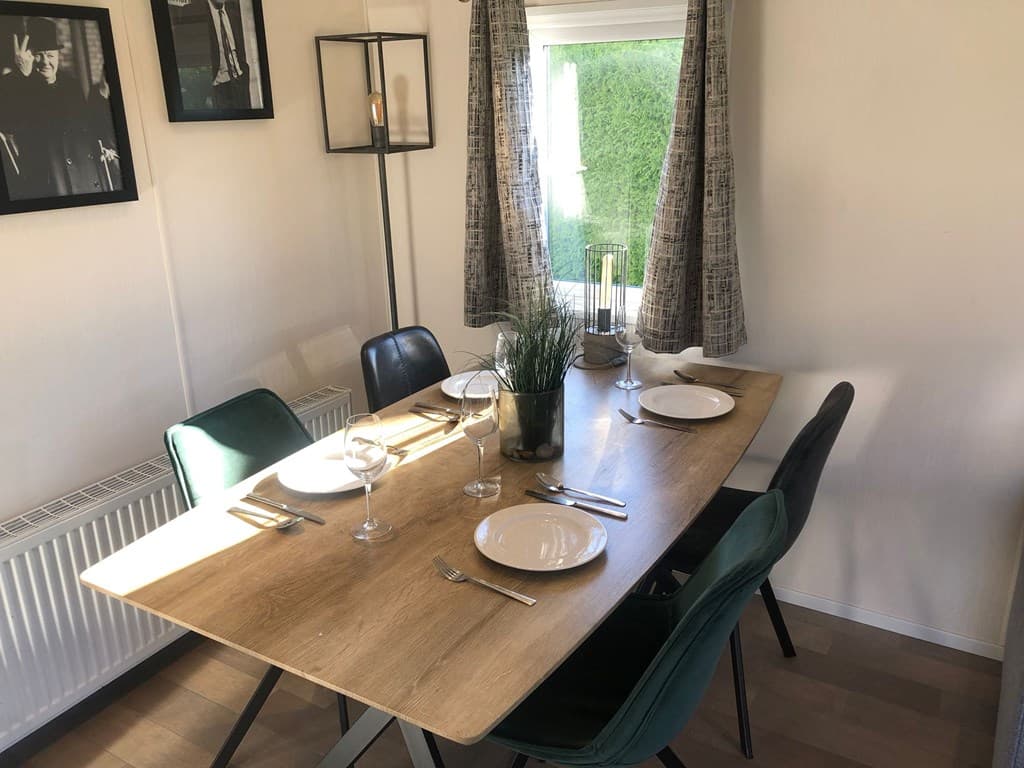 Eetkamertafel met glazen, borden en stoelen, raam met gordijnen, omringd door foto's aan de wand.