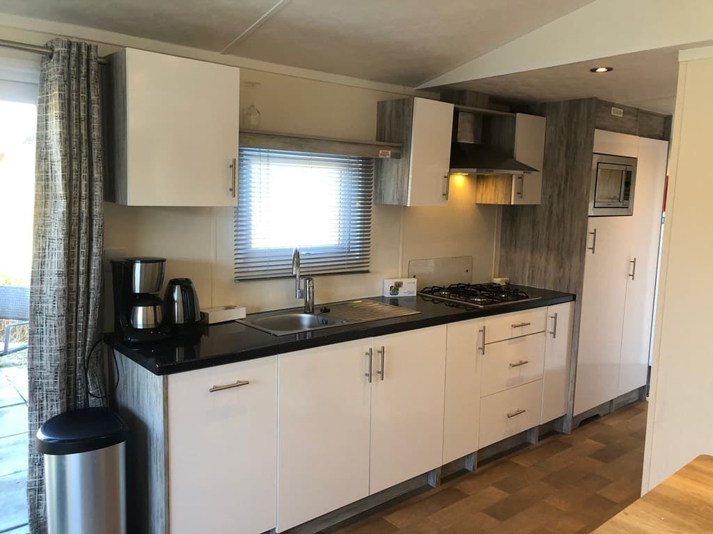 Moderne keuken met witte kasten, zwart werkblad, gasfornuis, koelkast en uitzicht op buitenruimte.