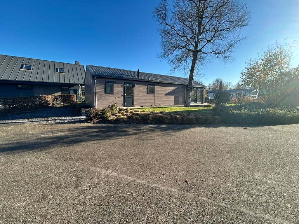 Moderne vakantiewoning met houtlook gevel en grote ramen, omgeven door groene tuinen en een parkeerplaats.