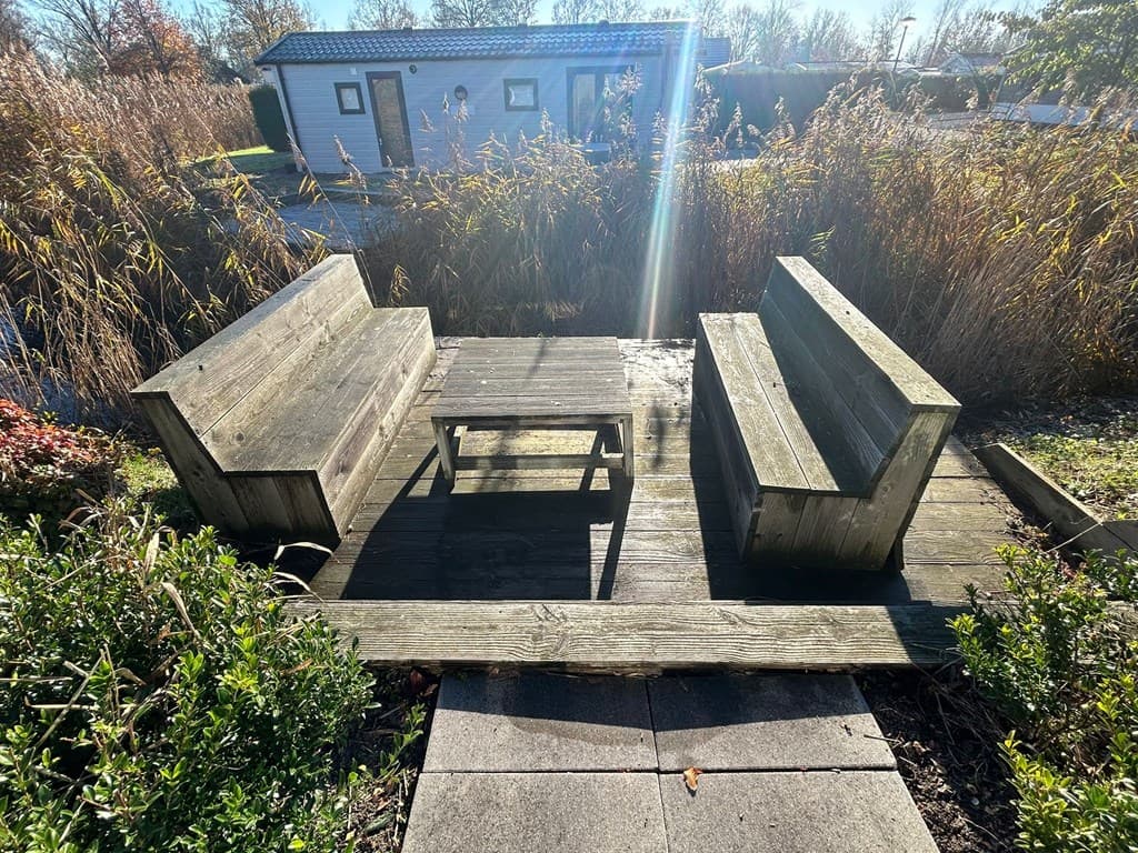 Houten zitplek met tafel, omringd door gras en riet, met een tweede woning op de achtergrond zichtbaar.