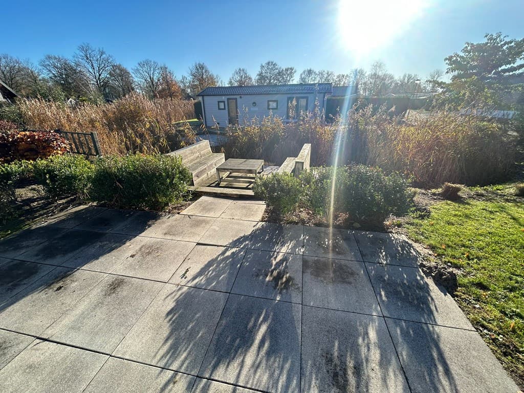 Tuin met terras en houten zitgedeelte omringd door groene struiken en gras, met een woning op de achtergrond.