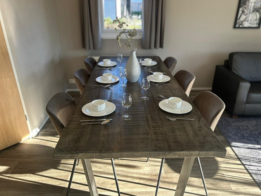 Eettafel met zes plaatsen, verzorgd gedekte borden en glazen, in een lichte woonruimte met raam en bank.