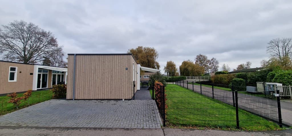 Buitenzicht van een vakantiewoning met houten gevel, groene grasvelden en een oprijlaan met straat en hekken