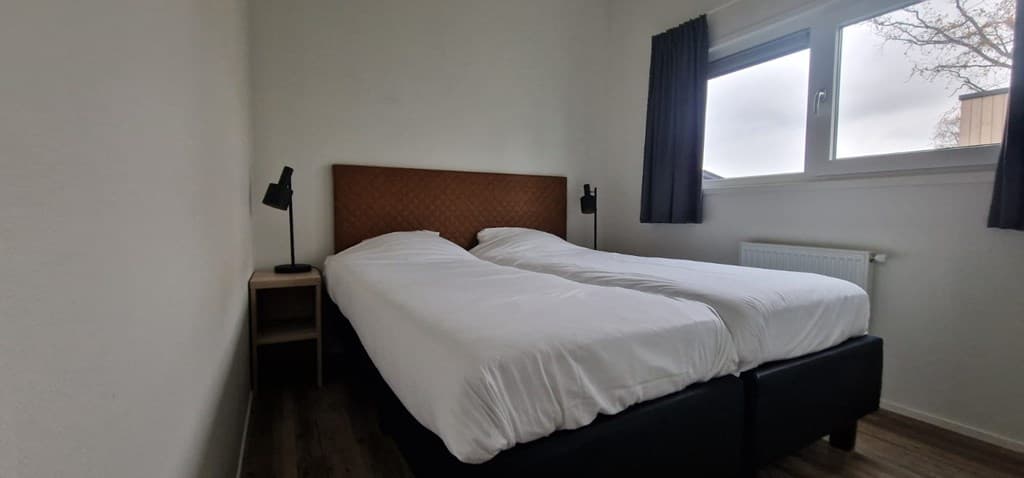 Slaapkamer met twee afzonderlijke bedden, nachtkastjes en een raam met gordijnen in een lichte ruimte.