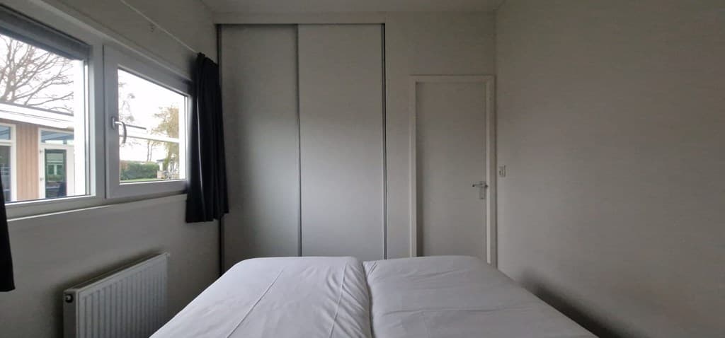 Slaapkamer met twee eenpersoonsbedden, witte muren en een inbouwkast, lichtinval via ramen aan de zijkant.