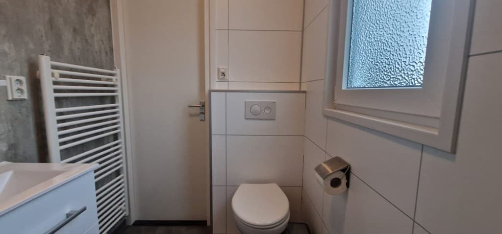 Badkamer met witte wandtegels, toilet, handdoekenradiator en venster met geribbeld glas
