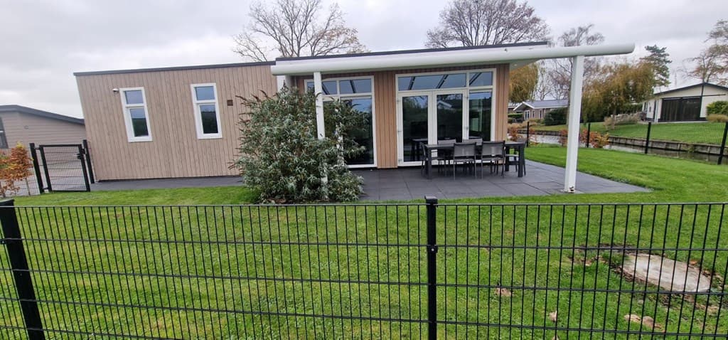 Moderne vakantiewoning met grote ramen, terras met zithoek en groene tuin, omringd door een hek.