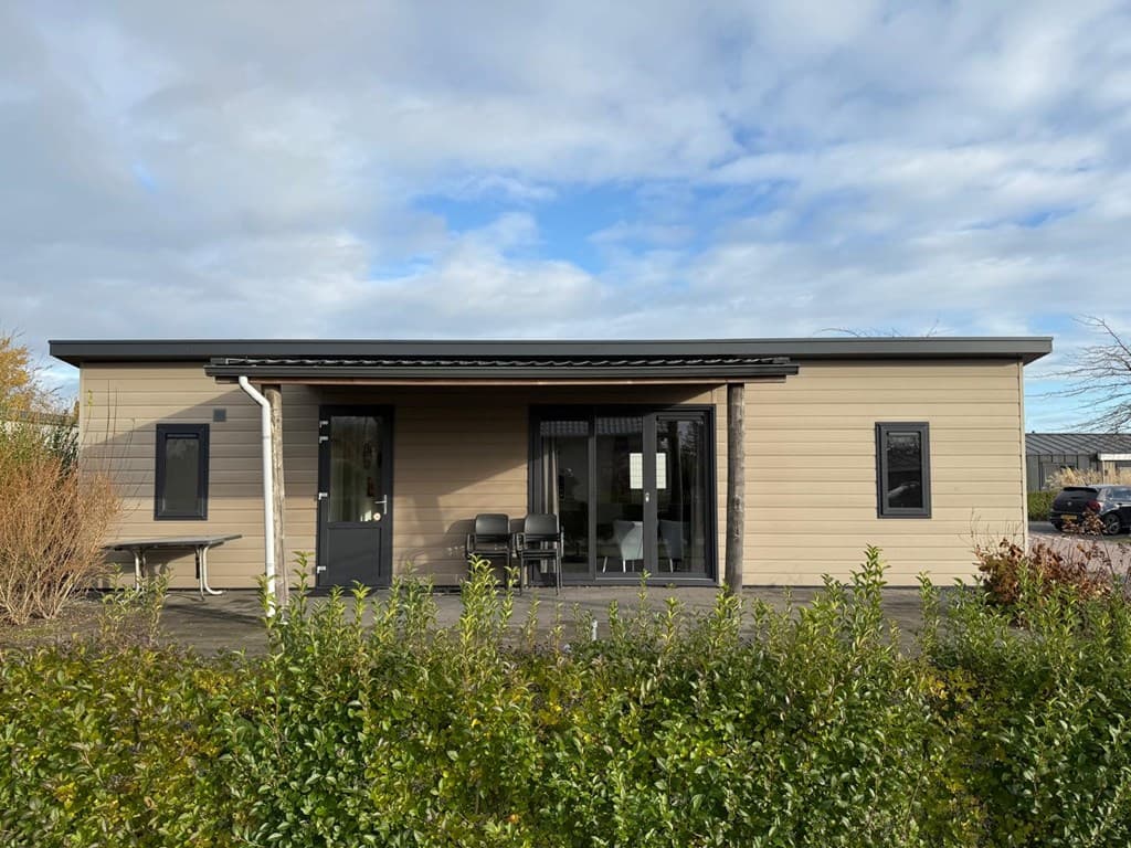Moderne vakantiewoning met een lichtbruine gevel, grote ramen en een terras aan de voorzijde, omgeven door groen.