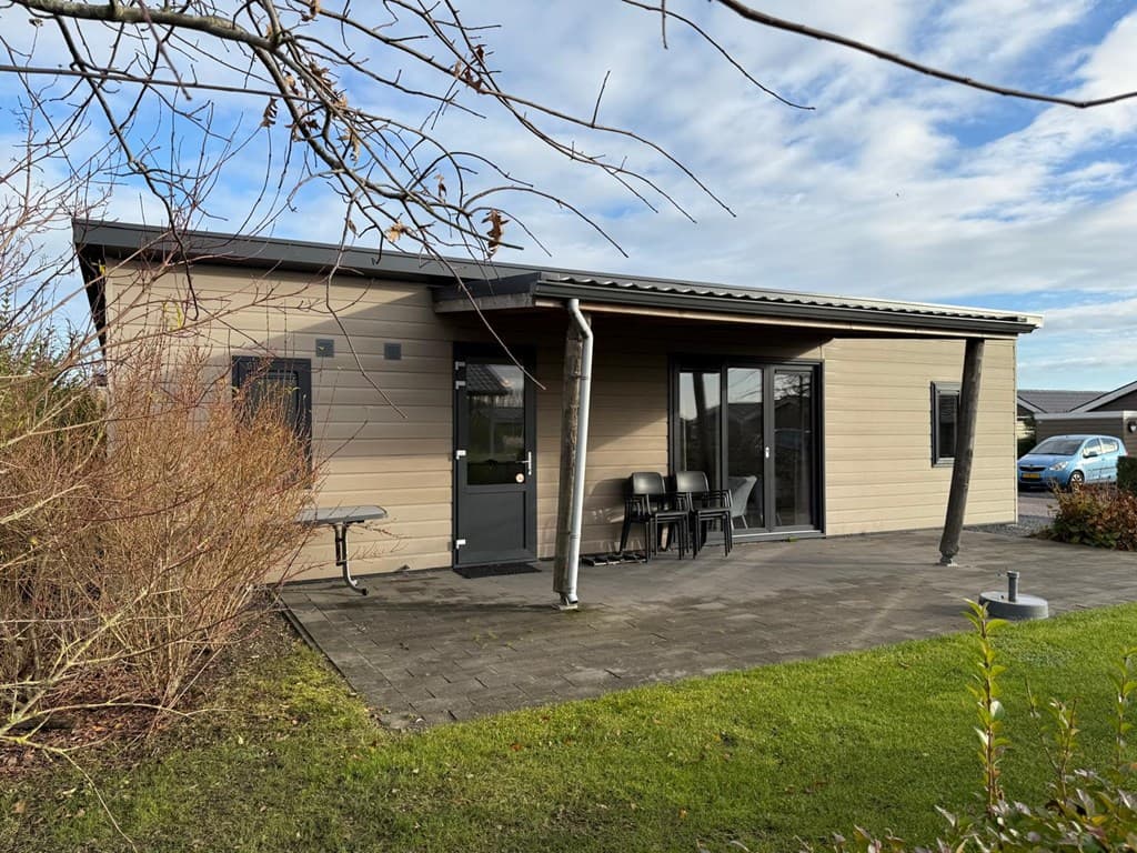Moderne vakantiewoning met ruime veranda, tafel en stoelen, omgeven door gras en struiken.
