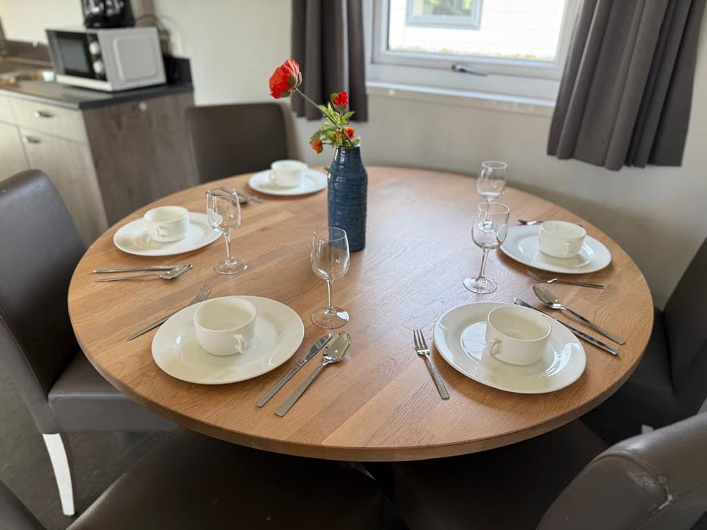 Eettafel met zes borden, glazen en bestek, met een vaas met bloemen op de tafel en een keuken op de achtergrond.