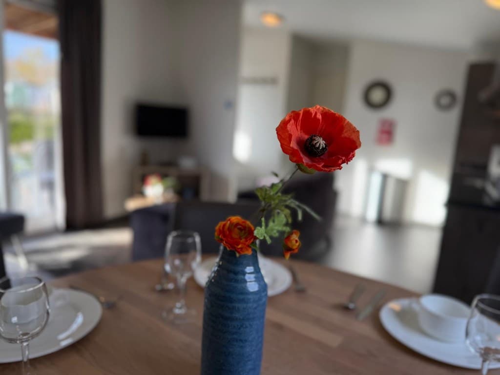 Eetkamer met tafel, servies en een vaas met bloemen, uitzicht op de woonkamer en keukenartiekelen zichtbaar op de achtergrond