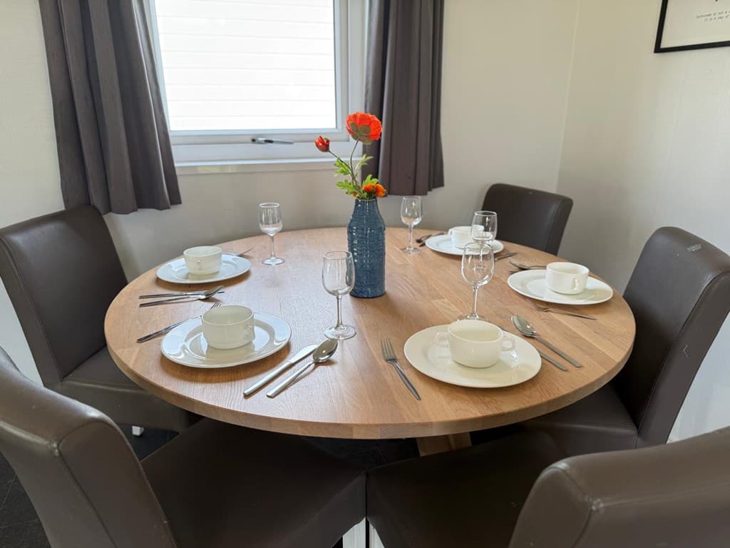 Eetkamer met ronde houten tafel, vier stoelen en gedekte tafel met borden, glazen en een vaas met bloemen.