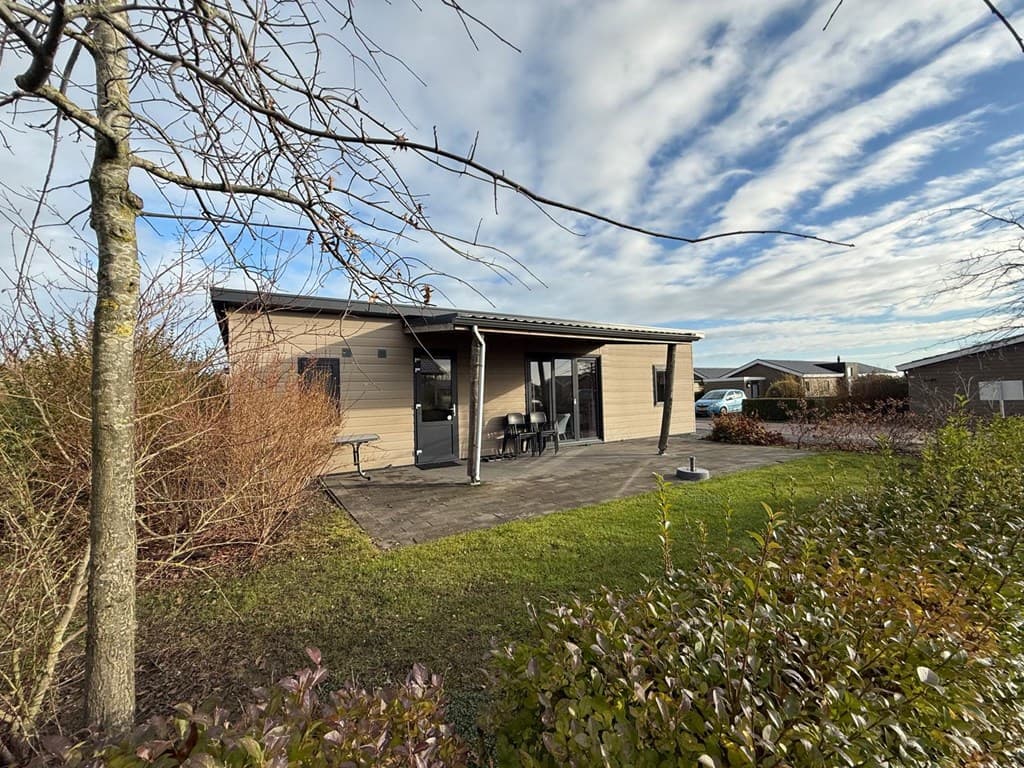 Vrijstaande woning met terras, omringd door gras en struiken, tegen een achtergrond van een heldere lucht en andere woningen.
