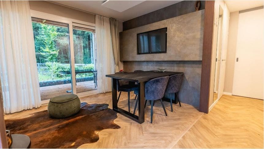 Eet- en woonkamer met houten vloer, modern tafeltje, vier stoelen en een tv, zicht op het terras met uitzicht op de natuur.
