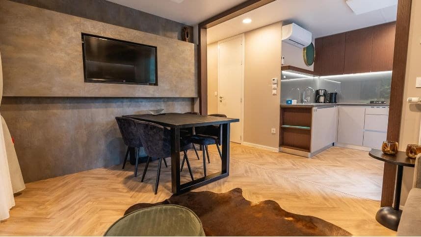 Moderne woonkamer met een tafel en stoelen, open keuken met koelkast en oven, en een televisie aan de muur.