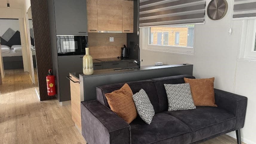 Moderne woonkamer met een bank, open keuken, en toegang naar een slaapkamer met inbouwbedden.
