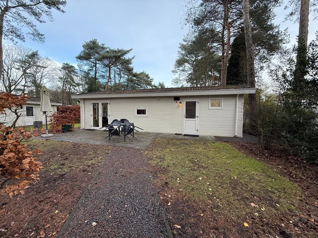 Buitenzicht van een lichte vakantiewoning met een betegeld terras en tuinmeubilair, omringd door bomen.