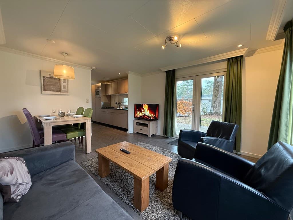 Zithoek met bank, fauteuils en salontafel, eetgedeelte met stoelen en open keuken, grote ramen met uitzicht op buiten.