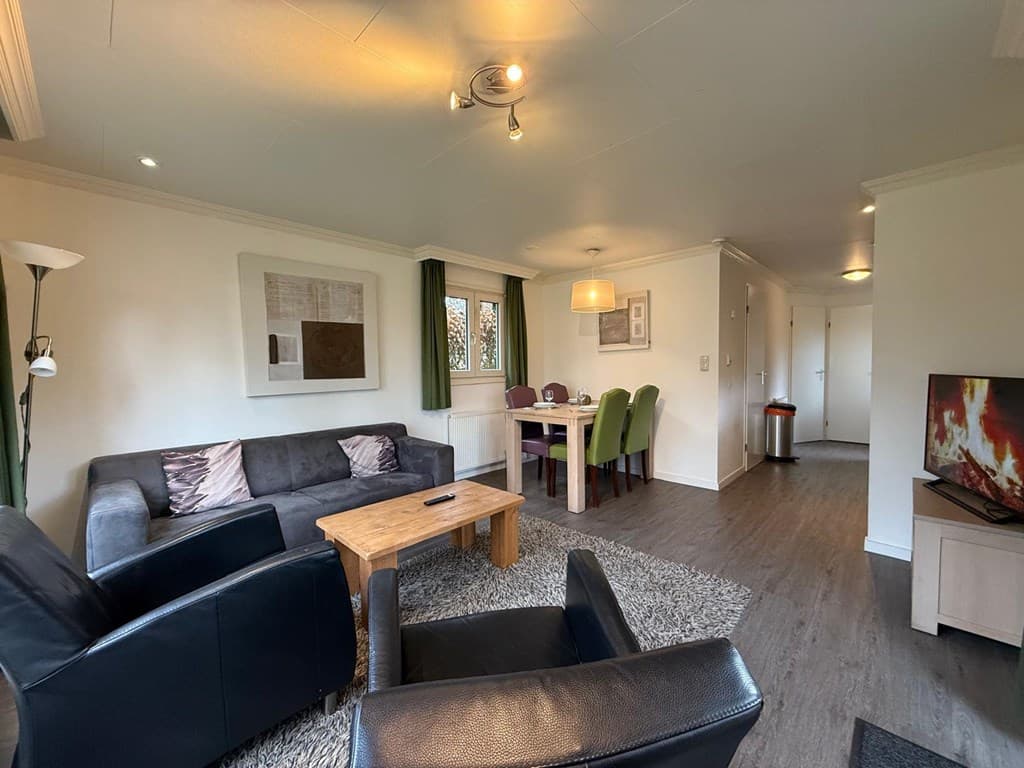 Gezellige woonkamer met bank, fauteuils, houten tafel en eethoek met groene stoelen in een lichte ruimte.
