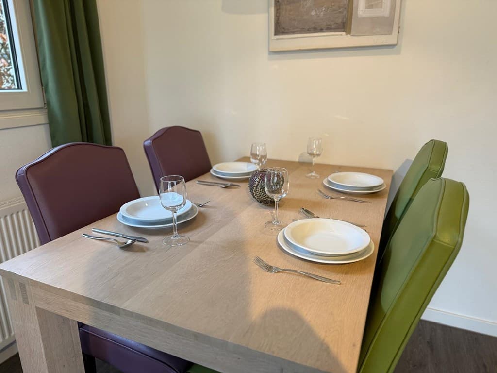 Eetkamertafel met vier stoelen, gedekt met bordjes, glazen en bestek in een lichte ruimte met groene en paarse accenten