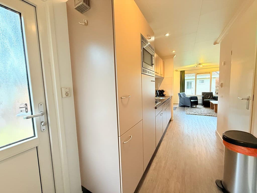 In een moderne vakantiewoning met een ruime gang, keuken aan de rechterkant en zicht op woonkamer met bank en lichtinval.