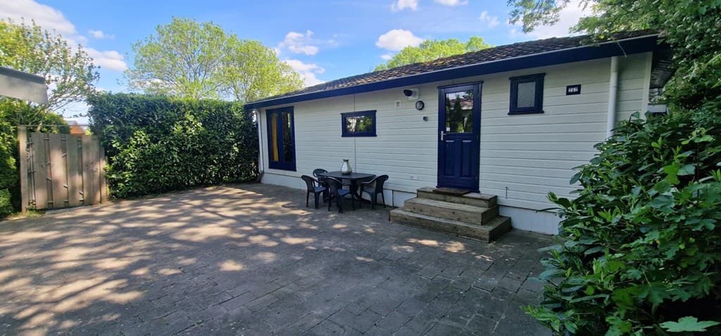Huisje met een houten terras, enkele meubels en omgeven door groene struiken en bomen onder een blauwe lucht