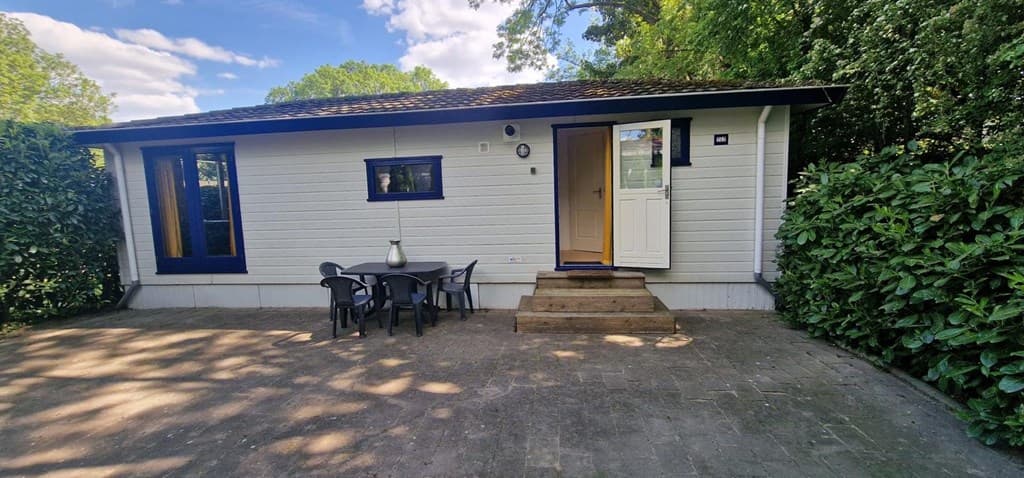 Witte vakantiewoning met een voordeur en terras met tafel en stoelen, omgeven door groen.