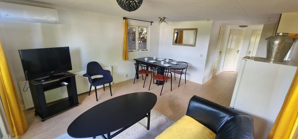 Woonkamer met een eettafel, zwarte bank, tv en moderne inrichting met gele gordijnen