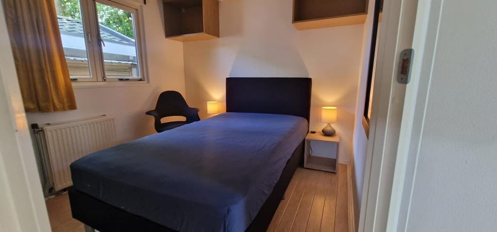 Slaapkamer met een enkel bed, nachtkastjes en een stoel naast het raam met gele gordijnen.