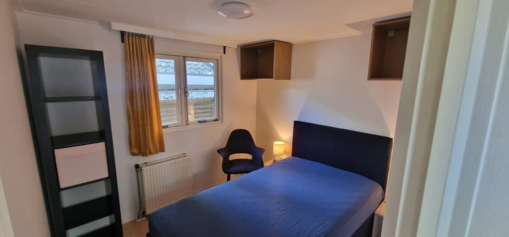 Slaapkamer met een bed, een stoel, een nachtkastje en een boekenkastje met openkasten en een raam met uitzicht naar buiten