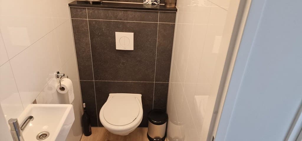 Kleine badkamer met toilet, wastafel en zwarte wandtegels, voorzien van een toiletpapierhouder en prullenbak