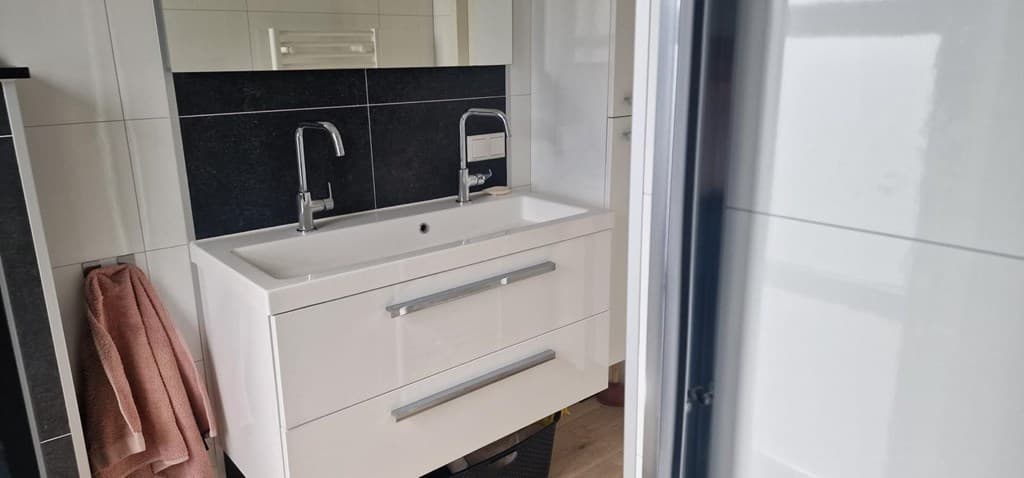Witte wastafel met dubbele kranen, zwarte wandtegels en een spiegel in een moderne badkamer.