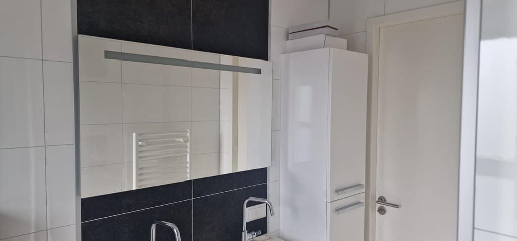 Moderne badkamer met een grote spiegel, wastafel en opbergkast tegen een wand van witte en donkere tegels.