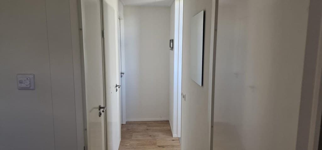 Een smalle gang met witte muren en houten vloer, deuren aan beide zijden en een spiegel aan de muur.