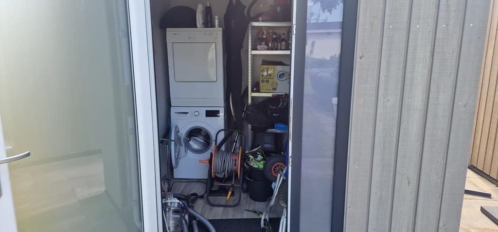 Ingang van een berging met een wasmachine, droger en verschillende huishoudelijke items zichtbaar.