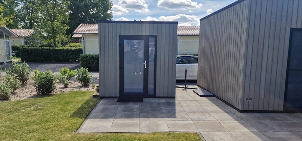 Zicht op een moderne houten vakantiewoning met een betegeld terras en groen plantsoen in een parkachtige omgeving