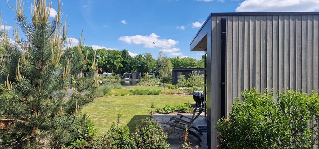 Dak en gevel van een moderne woning met uitzicht op een groene tuin en bomen onder een blauwe lucht met wolken
